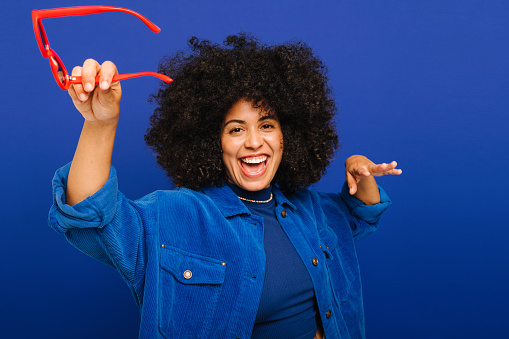 Happy young woman dancing and smiling cheerfully while standing against a blue background. Fashionable woman with curly hair celebrating and having a good time in a studio.