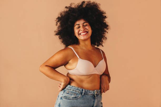 Curvy woman smiling happily while wearing a bra and jeans Self-confident young woman smiling happily while wearing a bra and jeans in a studio. Body positive young woman with Afro hair embracing her natural body and curves. bra stock pictures, royalty-free photos & images