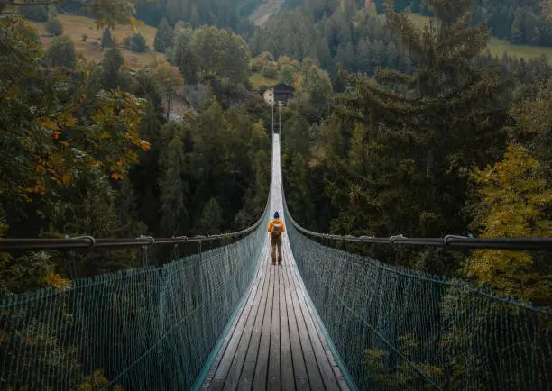 Photo of Young traveler man dressed in yellow jacket crosses hiking on an impressive wooden and metal bridge in the village of Goms in the Swiss alps