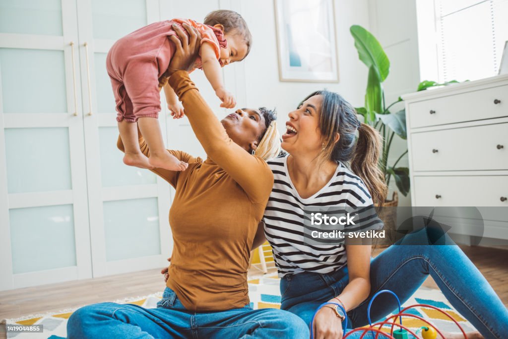 Gay couple with baby at home Young mothers playing with baby girl in living room. Lesbian Stock Photo