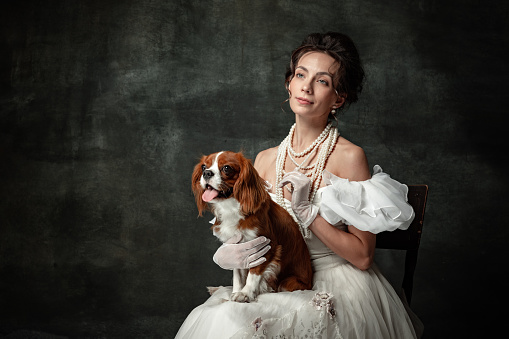 Lady with dog. Vintage portrait of young elegant woman in image of medieval person in renaissance style dress isolated on dark background. Comparison of eras, beauty, history, art, creativity.