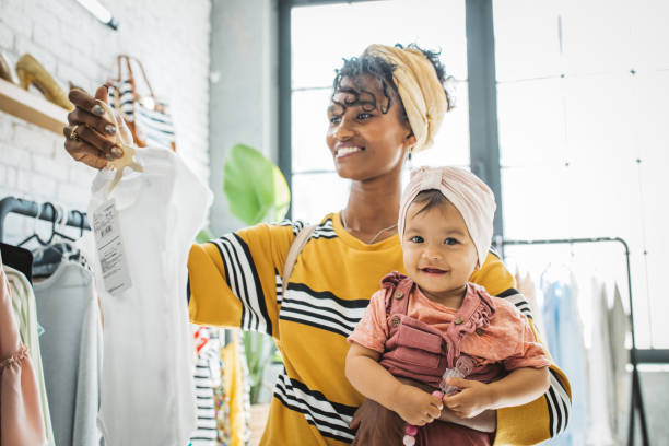 Sustainable shopping with baby Young woman with baby girl at thrift store, choosing clothes. baby boutique stock pictures, royalty-free photos & images