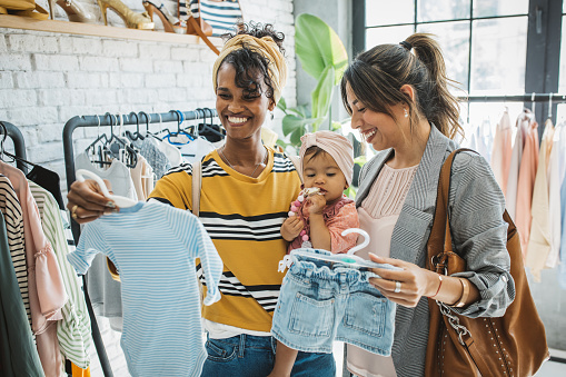 Young female gay couple at thrift store with baby girl, talking and choosing clothes.