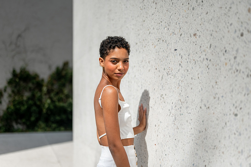 Beautiful young multiracial woman with short hair and white crop top posing by the concrete wall outdoors on a sunny day.