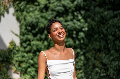 Beautiful young multiracial woman standing near the hedge and smiling with eyes closed.