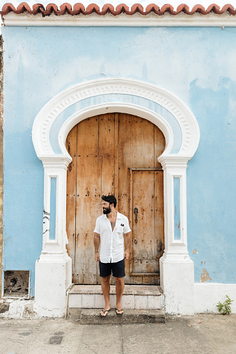 Man standing on a sidewalk with an old door behind him, he is posing for a photo. He is in a tourist town on vacation. This man is wearing a white shirt and shorts.