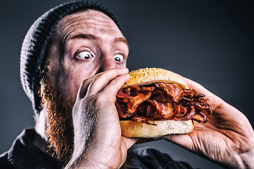 Studio photo of a burly red-haired, bearded man about to devour a sandwich made purely of fried bacon; eyes wide