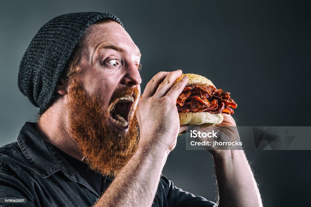 Burly Bearded Man Devouring a Bacon Sandwich Studio photo of a burly red-haired, bearded man about to devour a sandwich made purely of bacon; eyes wide and mouth agape Over Eating Stock Photo