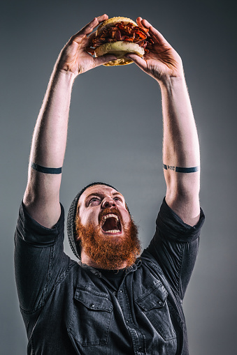Photo of a burly red-haired man holding a bacon sandwich over his head, screaming in victory as if it is a prized possession