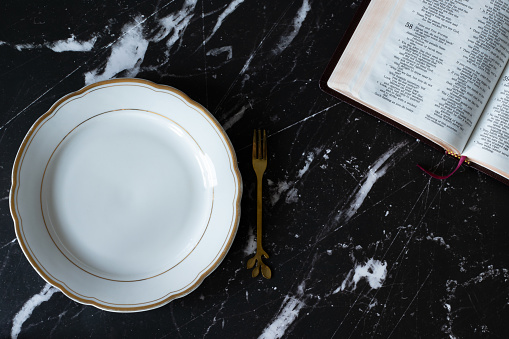 Empty plate, fork, and an open Holy Bible Book on a dark granite background. Top view. Fasting and prayer, Atonement Day, and repentance Christian biblical concept.