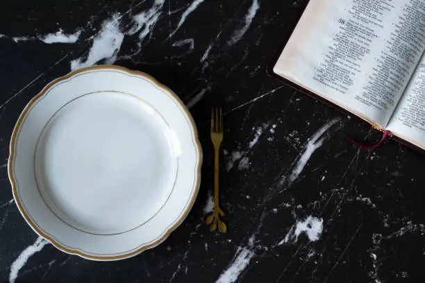 Empty plate, fork, and an open Holy Bible Book on a dark granite background. Top view. Fasting and prayer, Atonement Day, and repentance Christian biblical concept.