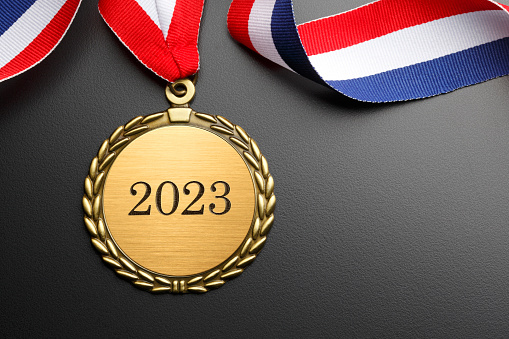 A gold medal engraved with 2023.  A red, white, and blue ribbon is attached to the medal that rests on a background that graduates from gray to black.