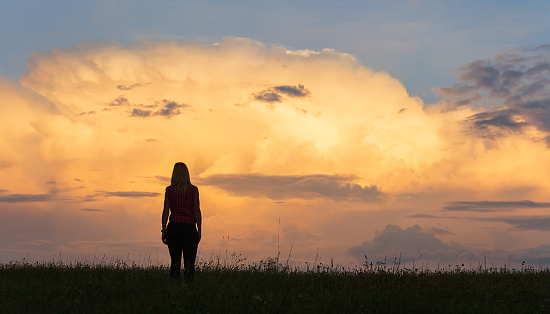 Silhouette of young caucasian woman in shirt walking through grass against sunset cloud sky