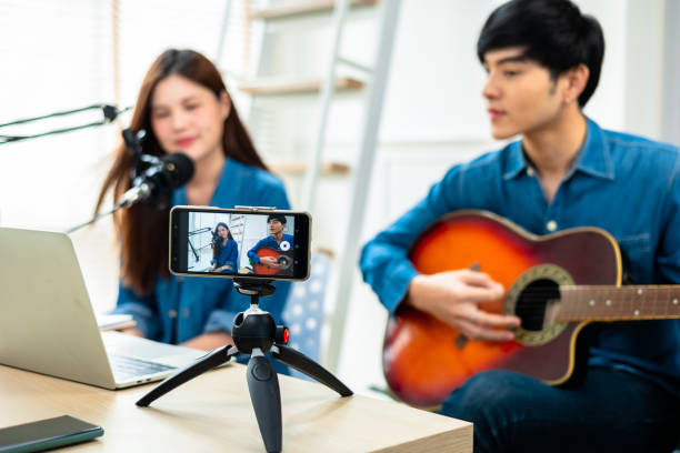istock Asian guitarist livestream virtual video tutorial at home,artist musician blogger sit on sofa in the living room . Happy young man singer use music instrument, compose song on cellphone. 1419088409