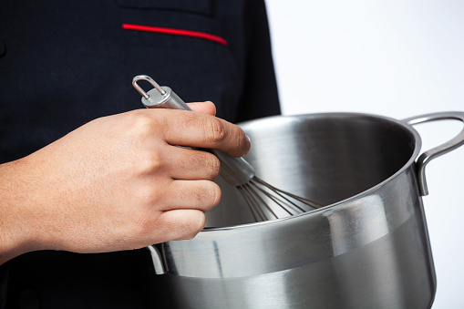 Closeup of cooks hand with metallic egg beater and pan.