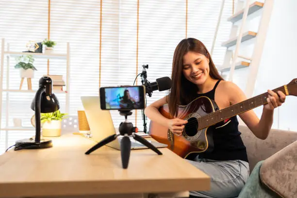 Asian guitarist livestream virtual video tutorial at home,artist musician blogger sit on sofa in the living room . Happy young man singer use music instrument, compose song on cellphone.