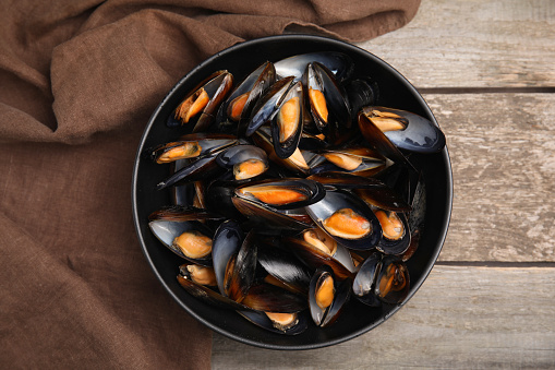 Bowl of delicious cooked mussels and napkin on wooden table, top view