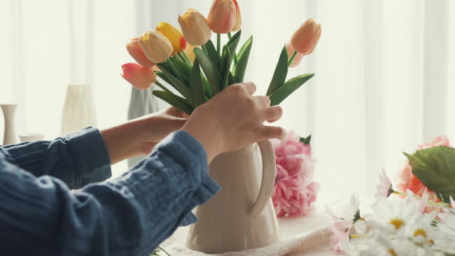 Close up of young Asian woman putting fresh colorful tulips into vase in the morning at home.