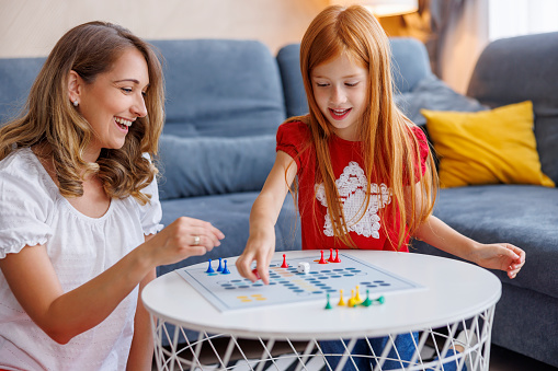 Beautiful mother and daughter having fun playing ludo board game while spending leisure time together at home