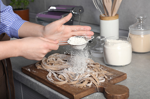 Woman sprinkling flour over uncooked soba at table in kitchen, closeup