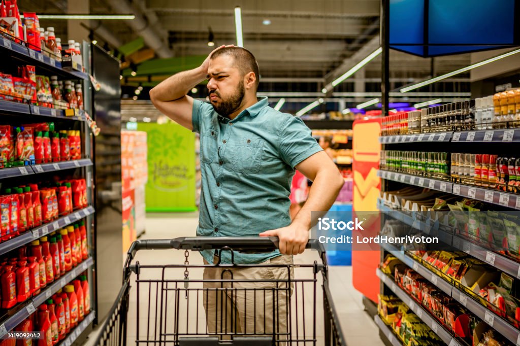 Man feeling upset about the increase in food prices Young man standing among the produce aisle at the supermarket and feeling upset about the increase in food prices Customer Stock Photo