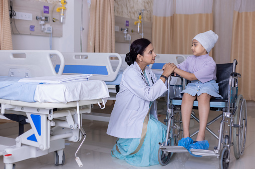 Female doctor talking to recovering girl patient sitting on wheelchair. Smiling child is looking at medical professional in hospital ward. They are at healthcare center.