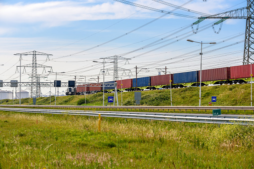 Cargo train loaded with containers running along a motorway in a port area on a clear summer day. Electricity pylons and fuel tanks are in background. Port of Rotterdam.
