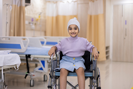 Portrait of sick girl sitting on wheelchair by beds. Ill little child wearing casuals undergoing treatment at ward in healthcare center. She is in hospital.