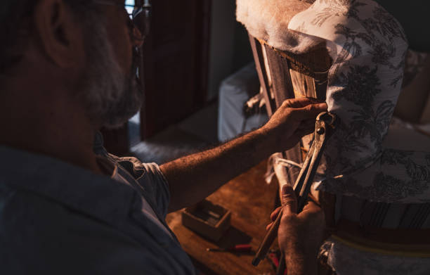Mature craftsman engaged in repair of vintage furniture, reupholstering chair in living room