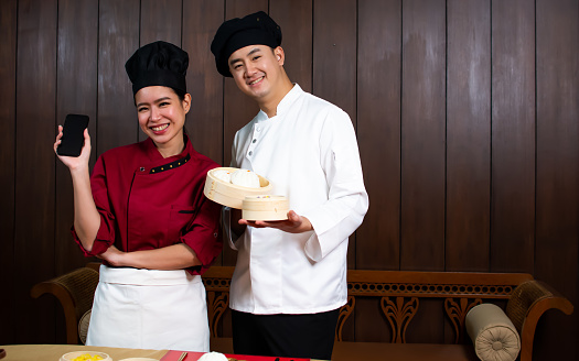 Two adult traditional professional couple Chinese chefs wearing uniform, hat, happily smiling, standing, presenting basket, mobile application for online food order, payment. Restaurant, Hotel Concept