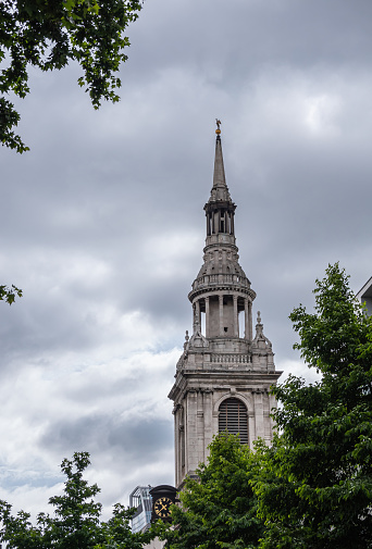 London, England, UK - July 6, 2022: Gray stone St Mary-le-Bow Church spire against gray cloudscape with green foliage on three sides.