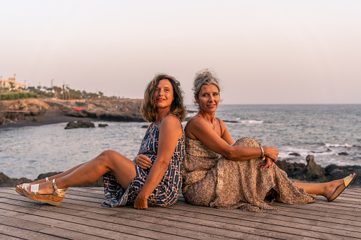 two lovely middle aged girlfriends sitting on a wooden bridge by the sea at sunset - summer vacation concept