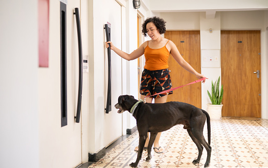 Smiling woman standing at an apartment elevator with her dog on a leash before going for a walk together