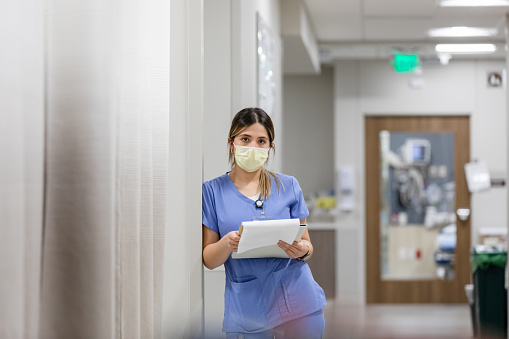 The female surgeon leans on the wall and looks at the camera while holding patient paperwork.