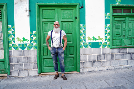 Full length photo of fashionable smiling senior man, positive emotions. Hipster guy wearing jeans and suspenders posing outdoor on the green door.