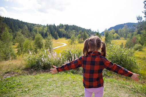 Caucasian child girl, back view, with open arms in a natural mountain landscape on a sunny day. Pian di Gembro natural park, Valtellina, Italy.