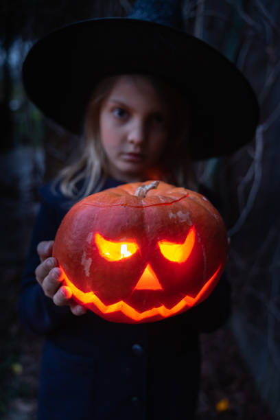 Girl in witch costume, hat with jack-o-lantern.Hand made from big pumpkin. Candle lights in eyes,nose,mouth. Celebratiion of halloween holiday.Cut by knife.Outdoors activity,backyard.Children's party Girl in witch costume, hat with jack-o-lantern.Hand made from big pumpkin. Candle lights in eyes,nose,mouth. Celebratiion of halloween holiday.Cut by knife.Outdoors activity,backyard.Children's party. halloween pumpkin human face candlelight stock pictures, royalty-free photos & images