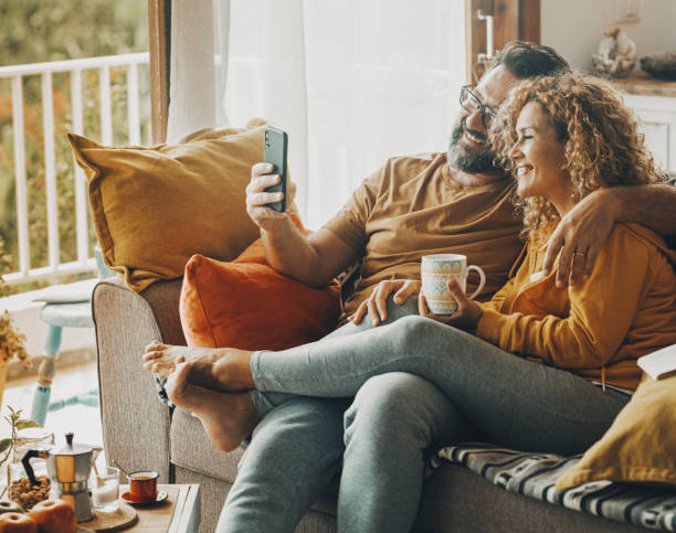 happy adult couple at home enjoy mobile phone connection doing video call conference with friends or parents away. technology lifestyle indoor house - comfortable relaxation sofa men imagens e fotografias de stock