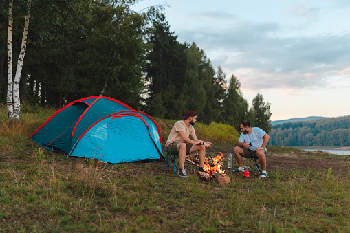Two young hikers camping outdoors. They have set up a tent and are talking by the campfire.