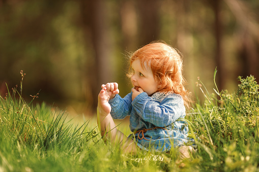 Funny red hair baby is sitting on the grass and holding her own leg like yoga. Cute child putting her feet up and showing new skills and development, the other hand is in the mouth. Baby stretching