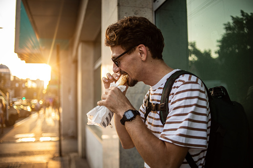 Spontaneous portrait of a handsome young man walking on a city street and eating a sandwich, a tourist in a big city.