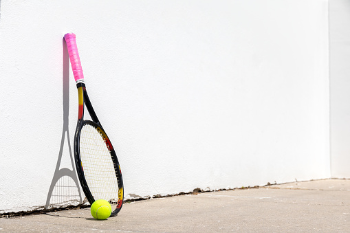 Tennis racquet and ball leaning against a white handball court wall. The wall is used for tennis practice through repetition.