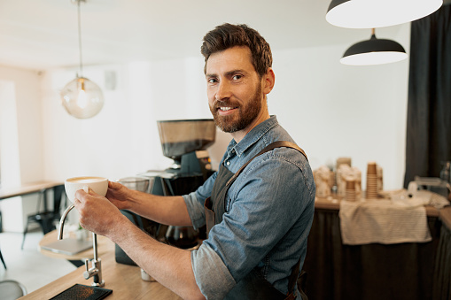 Handsome barista holding cup of coffee and look at the camera standing behind a counter