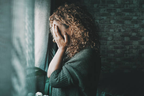 ritratto di lato donna triste e preoccupata a casa al buio guardando fuori dalla finestra. femmina preoccupata e sola sola in casa. concetto di fine della relazione ed emozione infruttuosa. signora che tocca il viso - tensione foto e immagini stock