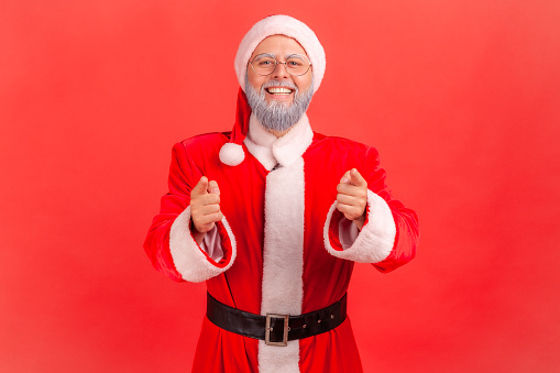 Smiling elderly man with gray beard wearing santa claus costume noticing and pointing finger to camera, choosing lucky winner, smiling joyfully. Indoor studio shot isolated on red background.