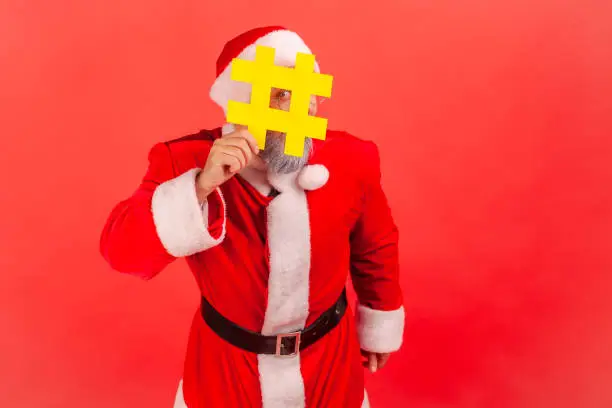 Photo of Portrait of playful elderly man in santa claus costume hiding face behind hashtag symbol, popular blogger recommending to follow tag, comments.