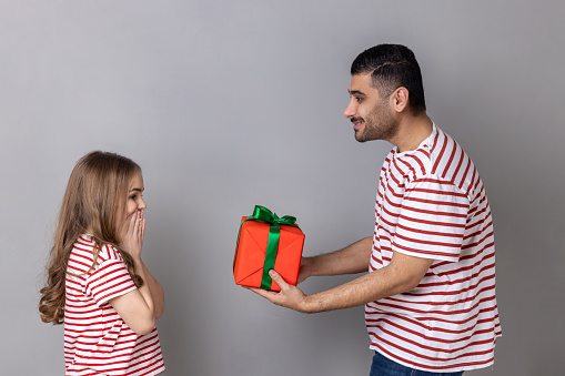 Side view of father giving red present box to his daughter, family in striped T-shirts, excited little girl being happy to get a gift on her birthday. Indoor studio shot isolated on gray background.