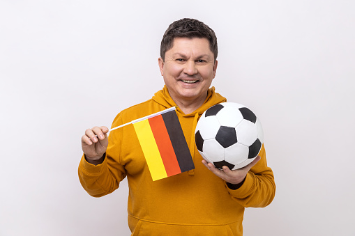 Middle aged man with toothy smile holding flag of germany and soccer black and white classic ball and watching match, wearing urban style hoodie. Indoor studio shot isolated on white background.