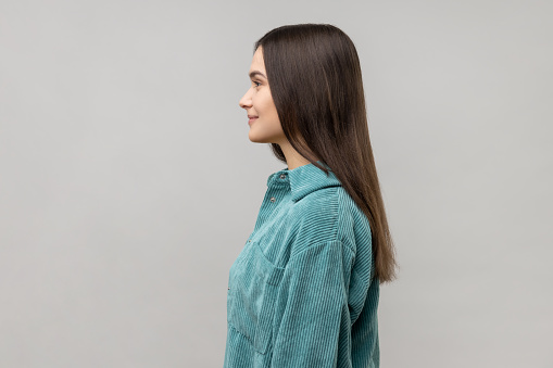 Side view of happy successful young woman standing looking at camera with charming and joyous toothy smile, wearing casual style jacket. Indoor studio shot isolated on gray background.