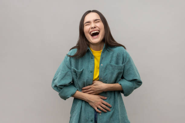 Happy woman holding her stomach and laughing out loud, chuckling giggling at amusing anecdote. Happy woman holding her stomach and laughing out loud, chuckling giggling at amusing anecdote, sincere emotion, wearing casual style jacket. Indoor studio shot isolated on gray background. belly laugh stock pictures, royalty-free photos & images